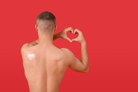 Young man with sunscreen cream on his body making heart gesture against red background, back viewの写真素材