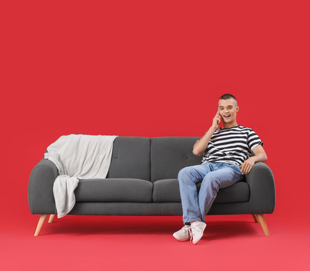 Happy young man sitting on sofa and talking by mobile phone against red backgroundの写真素材