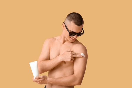Happy young man in sunglasses applying sunscreen cream on his body against orange backgroundの写真素材