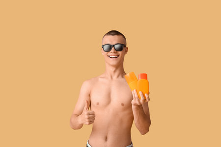 Happy young man in sunglasses with bottles of sunscreen cream showing thumb-up gesture on orange backgroundの写真素材