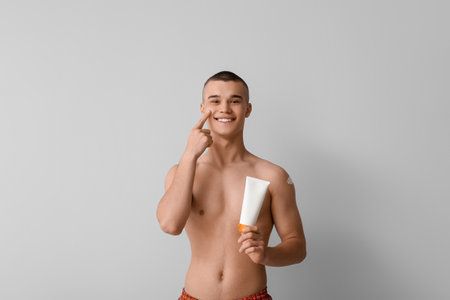 Happy young man applying sunscreen cream on his face against gray backgroundの写真素材