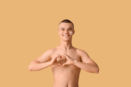 Happy young man with sunscreen cream on his face making heart gesture against orange backgroundの写真素材