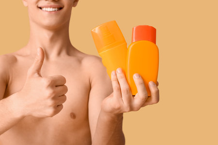 Happy young man with bottles of sunscreen cream showing thumb-up gesture on orange backgroundの写真素材