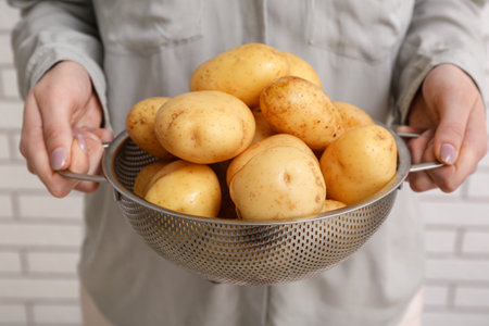 Woman holding colander with raw potatoes near white brick wallの写真素材