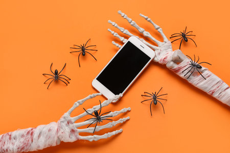 Skeleton hands with modern mobile phone and spiders for Halloween celebration on orange background
