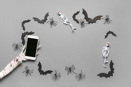 Frame made of skeleton hand, modern mobile phone, paper bats and spiders for Halloween celebration on gray backgroundの写真素材