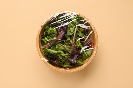 Bowl of fresh salad covered with plastic food wrap on beige background