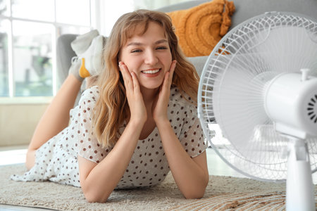 Beautiful happy young woman with electric fan lying on floor in living roomの写真素材