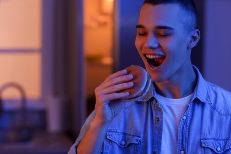 Hungry young man eating burger in kitchen at night, closeup