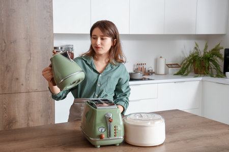 Young woman in kitchen with electric kettle, toaster and juicer