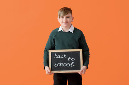 Little schoolboy in stylish uniform holding blackboard with text BACK TO SCHOOL on orange backgroundの写真素材