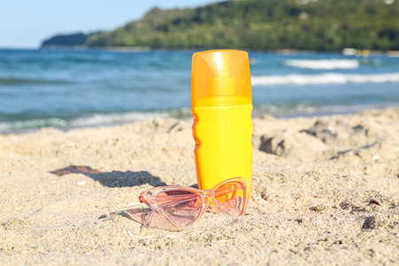 Bottle of sunscreen cream with sunglasses on sand near ocean at beach