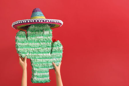 Female hands holding pinata in shape of cactus and sombrero on red background