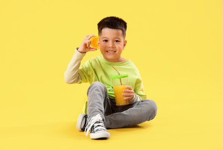 Little Asian boy with cup of fresh citrus juice and orange slice sitting on yellow backgroundの写真素材
