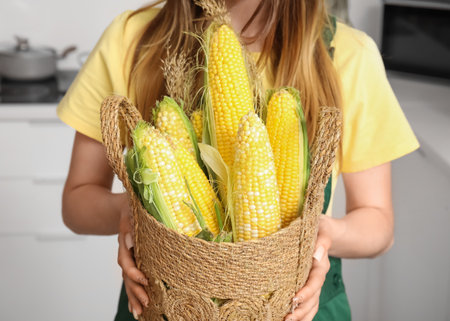 Beautiful young female farmer with wicker basket full of ripe corn cobs in kitchenの写真素材