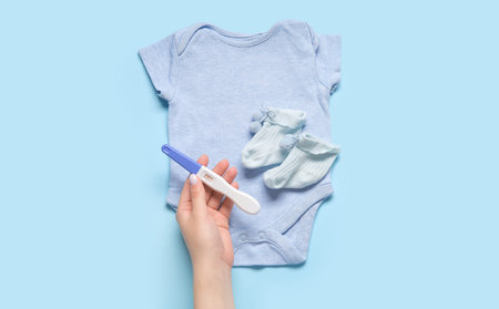 Female hand with pregnancy test and baby clothes on color background