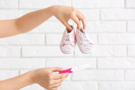 Female hands with positive pregnancy test and baby booties against light brick wall, closeupの写真素材
