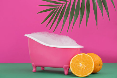 Small bathtub with soap foam, oranges and palm leaf on color backgroundの写真素材