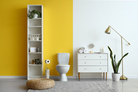 Interior of modern restroom with ceramic toilet bowl, shelving unit and chest of drawers near yellow wallの写真素材