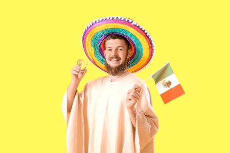 Handsome young man in sombrero with Mexican flag and shot of tequila on yellow backgroundの写真素材