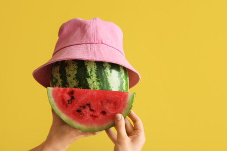 Female hands with whole ripe watermelon with slice and bucket hat on yellow backgroundの写真素材