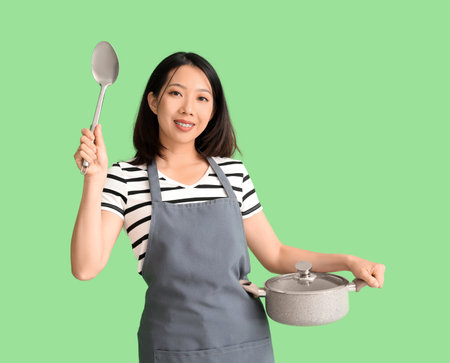 Happy beautiful Asian woman with ladle and saucepan on green backgroundの写真素材