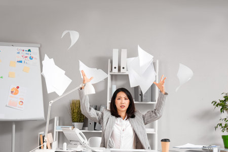 Stressed Asian businesswoman sitting at table and throwing paper in office