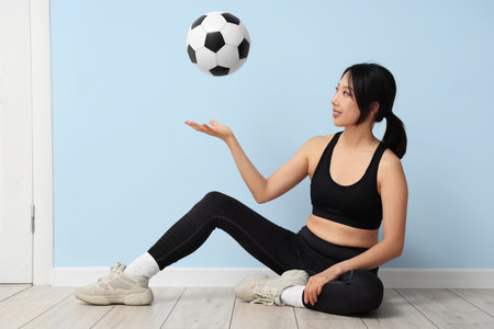 Happy young sporty Asian woman with soccer ball sitting on floor near blue wallの写真素材