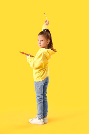 Cute little girl with palette paint and brush on yellow backgroundの写真素材