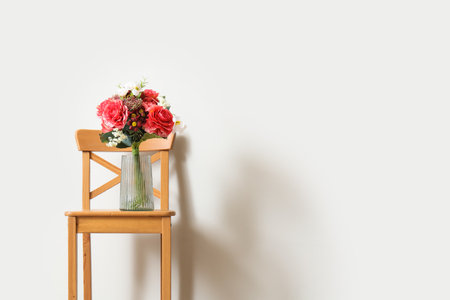 Vase with bouquet of beautiful artificial flowers on wooden chair near white wallの写真素材