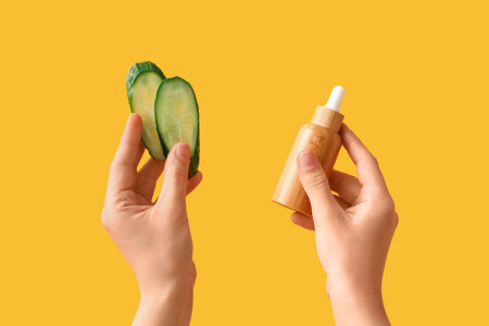 Woman with cosmetic dropper bottle and cucumber slices on yellow backgroundの写真素材