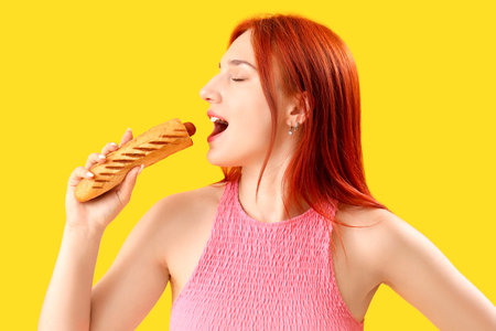 Beautiful young woman eating tasty French hot dog on yellow backgroundの写真素材