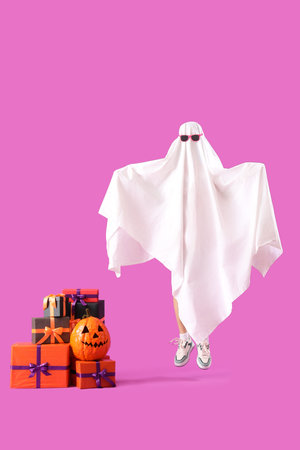 Young woman dressed as ghost with Halloween pumpkin and gifts on pink backgroundの写真素材