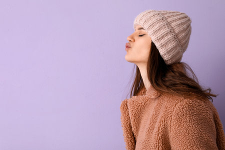 Young woman in teddy coat blowing kiss on lilac backgroundの写真素材