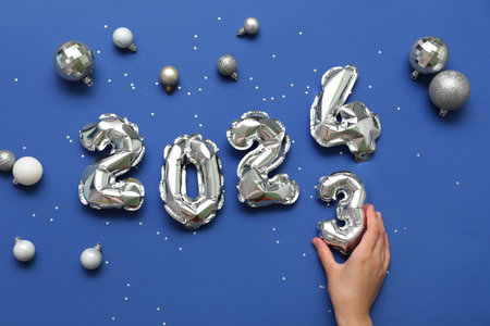 Female hand with figure 2024 made of balloons and Christmas decorations on blue backgroundの写真素材