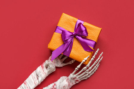 Gift box and skeleton hands for Halloween celebration on red backgroundの写真素材