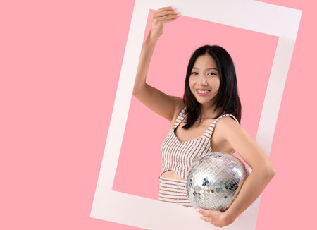 Young beautiful Asian woman with frame and disco ball on pink backgroundの写真素材