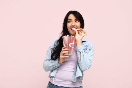 Happy young woman with bucket of popcorn on pink backgroundの写真素材