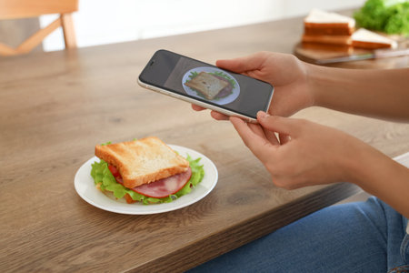 Young woman with mobile phone taking picture of tasty sandwich in kitchen, closeupの写真素材