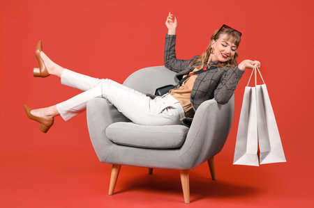 Stylish young woman with shopping bags in armchair on red backgroundの写真素材
