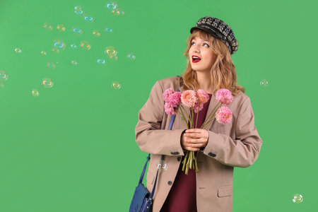 Stylish young woman in fall clothes with flowers and soap bubbles on green background