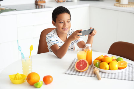 Little African-American boy with mobile phone, fresh juice and plate of different citruses in...