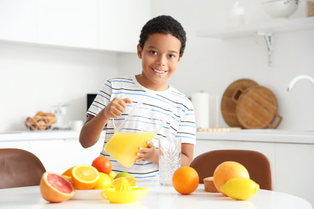 Little African-American boy pouring fresh citrus juice into glass from jug at table in kitchenの写真素材