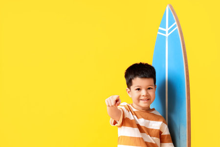 Cute little Asian boy with surfboard pointing at camera on yellow background