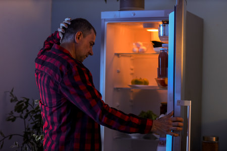 Hungry mature man near open fridge in kitchen at nightの写真素材