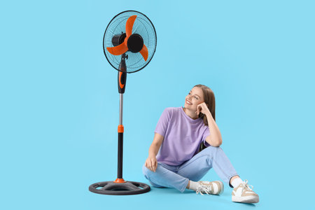Young woman with electric fan sitting on blue background