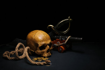 Human skull with sword, toy model of cannon, pirate hat and coins on black backgroundの写真素材