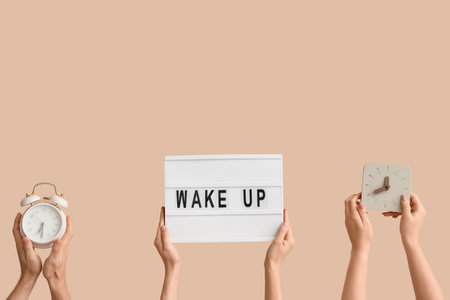 Hands holding alarm clocks and board with text WAKE UP on beige backgroundの写真素材