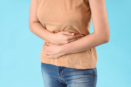 Young beautiful woman suffering from abdominal pain on blue background, closeup