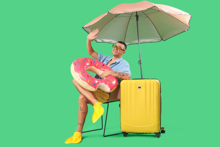 Happy young man with inflatable ring, umbrella and suitcase sitting on chair against green backgroundの写真素材